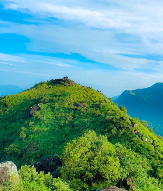 A panoramic view of rolling green hills from a high vantage point. The sky is a soft blue with scattered white clouds, creating a peaceful and expansive natural scene.