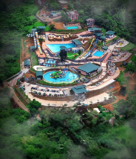 A modern water park facility located on a hillside. The pool is surrounded by palm trees and overlooks a vast valley, combining adventure with scenic beauty.